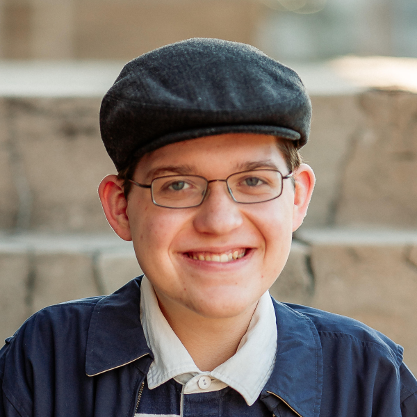 A person wearing glasses, a dark flat cap, and a blue jacket is smiling at the camera with stone steps in the background.