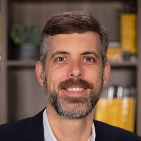 A man with short brown hair and a beard, wearing a dark jacket and light shirt, is smiling in front of a bookshelf filled with books.