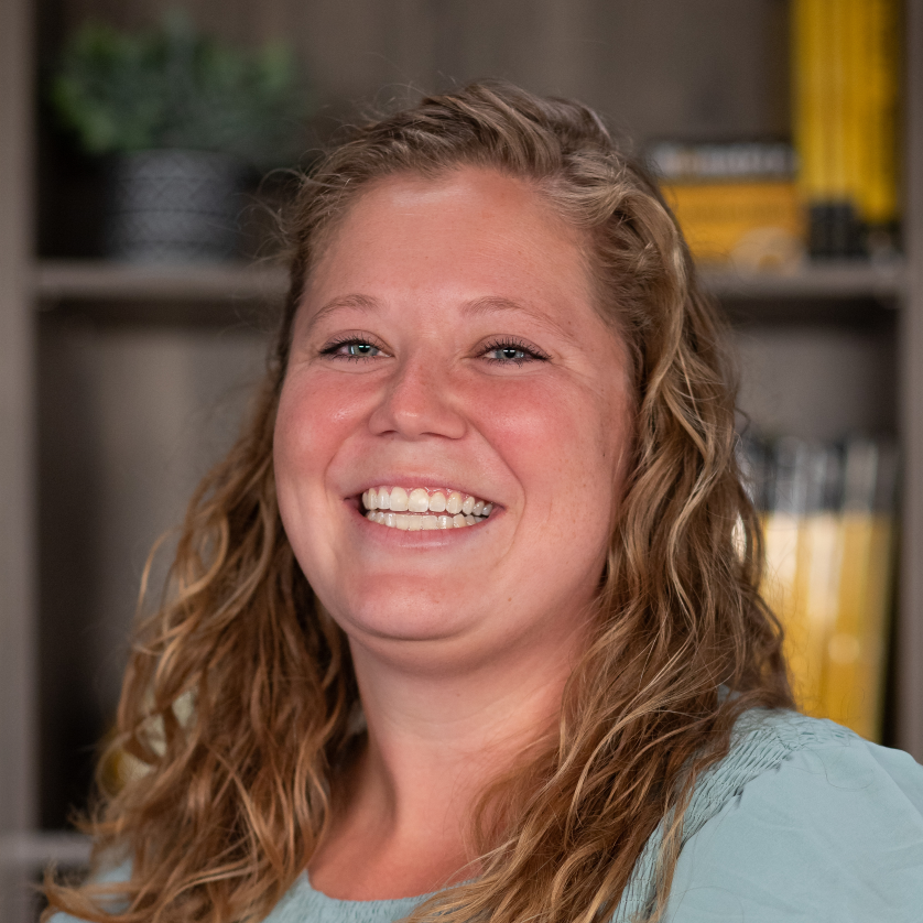 A woman with wavy blonde hair wearing a light blue top smiles at the camera. Shelves with books and a plant are visible in the blurred background.