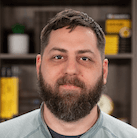 A bearded man with short dark hair stands indoors in front of a blurred bookshelf filled with books and decorative items.